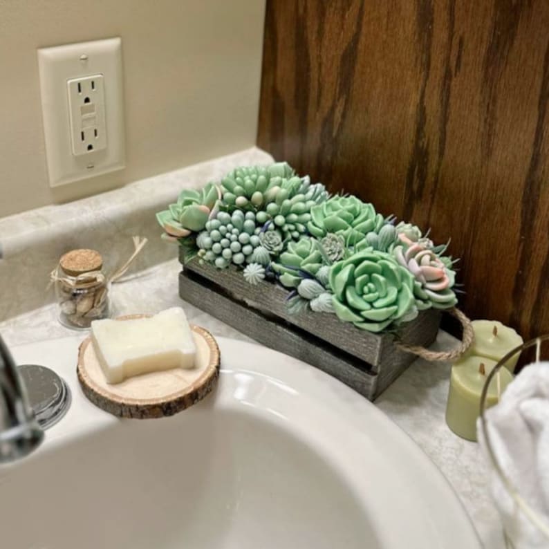 Puede incluir: Una caja de madera gris llena de una variedad de plantas suculentas verdes y rosadas. La caja est&aacute; sentada en una encimera de ba&ntilde;o blanca.