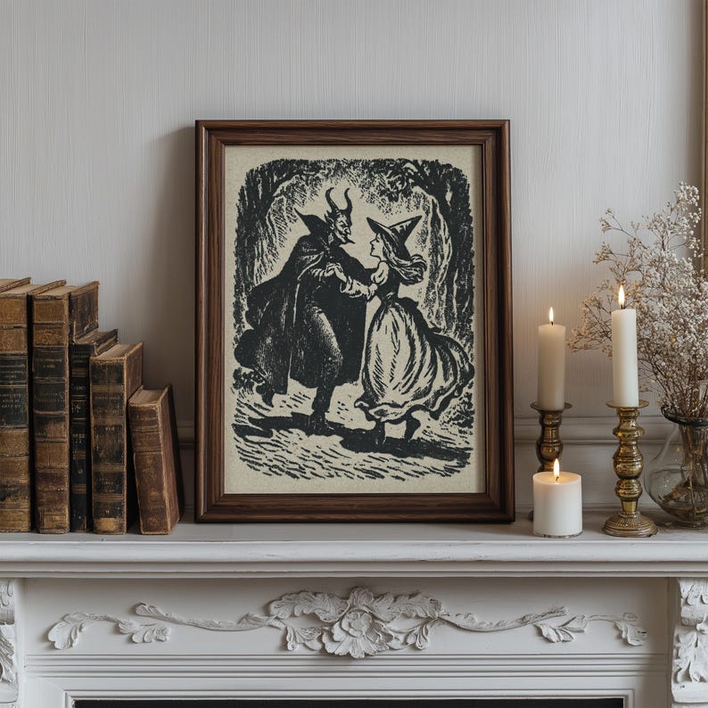 May include: A framed black and white print shows a devil and a witch dancing in a forest. The artwork is in a dark brown wooden frame. The scene is set on a white mantelpiece with lit candles and antique books.