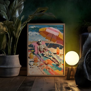 May include: A framed art print depicts a skeleton relaxing on a beach under a striped umbrella. The artwork features vibrant colors, including blue, pink, yellow, and orange. A cocktail and other beach items are also visible. The print is displayed on a wooden floor.