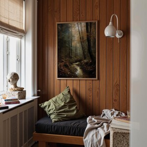 May include: A framed art print of a forest scene with a stream hangs on a wood-panelled wall. An olive green cushion and black cushion are below the artwork. A white wall sconce and a window with a beige blind are also visible.
