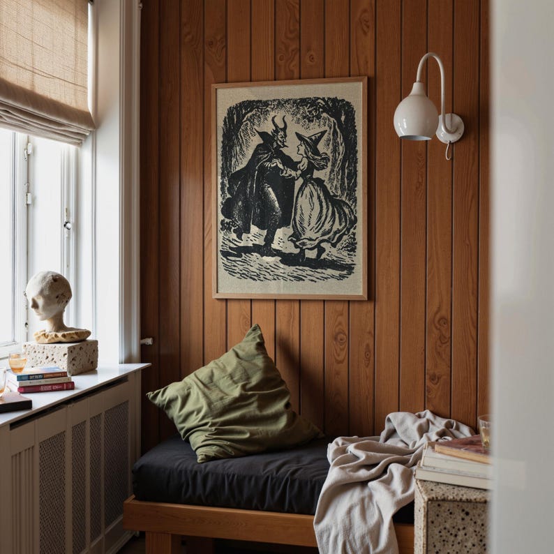 May include: A framed black and white print of a devil and a witch dancing in a forest, mounted on a wood-panelled wall. A green cushion sits on a black seat, and a white lamp is fixed to the wall. The room has a vintage, cosy feel.
