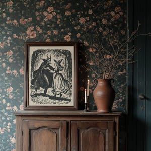 May include: A framed black and white print depicting a devil and a witch dancing in a forest setting. The print rests on a wooden cabinet, accompanied by a terracotta vase with branches and lit candles. Floral wallpaper forms the backdrop.