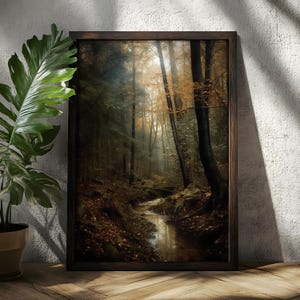 May include: A framed art print of a forest scene with a stream. The artwork uses a dark, moody colour palette with shades of brown, green, and gold. The stream winds through the forest, reflecting the light. The frame is dark brown.