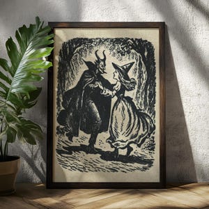 May include: Framed black and white print featuring a devil in a cloak dancing with a witch in a dress and hat. The artwork is set in a forest scene, with trees and foliage in the background. The frame is dark brown.