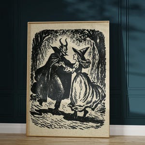 May include: A framed black and white vintage print depicting a devil and a witch dancing in a forest setting. The artwork is in a gold-coloured frame, set against a dark teal wall. The illustration style is reminiscent of classic Halloween imagery.