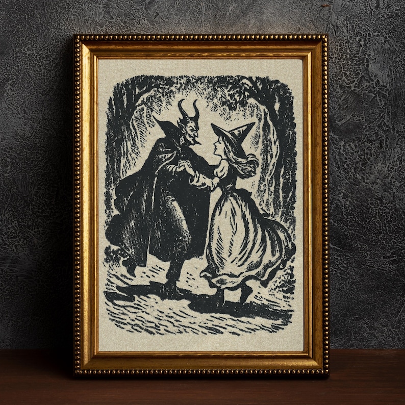 May include: A framed black and white illustration shows a devil and a witch dancing in a forest. The artwork is in a gold-coloured frame. The devil wears a cape and has horns. The witch wears a pointed hat and a long dress. The image has a vintage, Halloween theme.