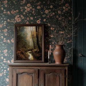 May include: A framed landscape painting of a forest scene with a stream, displayed on a wooden cabinet. A large terracotta vase with branches and two lit candles sit on the cabinet. The background features floral wallpaper.