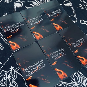 May include: A collection of tarot cards with black backgrounds and orange flame designs. Each card features white text with phrases like "I want to make things right" and "I'm scared you will reject me." The cards are arranged on a black cloth with white celestial patterns.