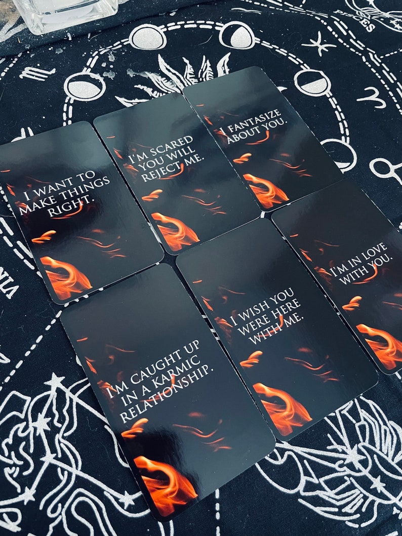 May include: A collection of tarot cards with black backgrounds and orange flame designs. Each card features white text with phrases like "I want to make things right" and "I'm scared you will reject me." The cards are arranged on a black cloth with white celestial patterns.
