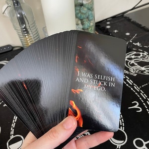 May include: A deck of black tarot cards fanned out, with the top card showing a fiery design and the text "I WAS SELFISH AND STUCK IN MY EGO." The cards are held in a hand, with a white background.