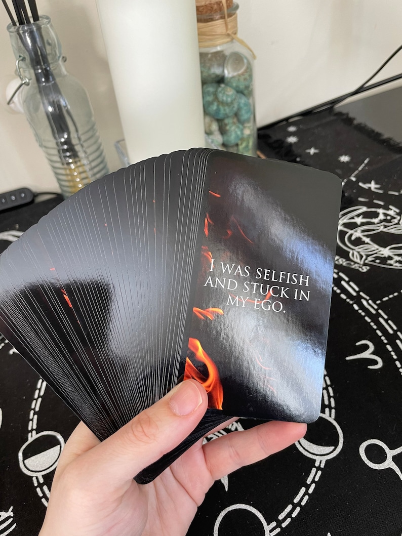 May include: A deck of black tarot cards fanned out, with the top card showing a fiery design and the text "I WAS SELFISH AND STUCK IN MY EGO." The cards are held in a hand, with a white background.