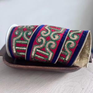 May include: A roll of decorative ribbon featuring a vibrant red velvet base with deep blue velvet borders. The ribbon is embellished with intricate, swirling patterns outlined in shimmering gold sequins and filled with bright green glitter accents.