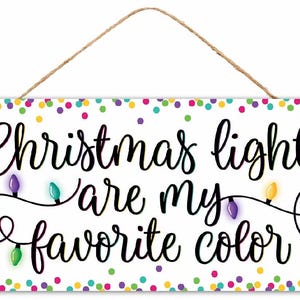 May include: Rectangular white sign with the text "Christmas lights are my favorite color" in black script. The sign features a string of colorful Christmas lights and a confetti border, with a jute rope for hanging.
