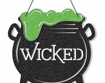 May include: A black cauldron-shaped sign with the word "WICKED" in white lettering. The cauldron has a glittery green bubbling potion at the top. The sign is suspended by two thin gray strings.