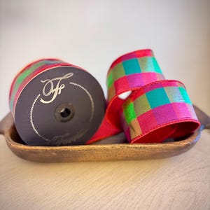 May include: A spool of ribbon featuring a vibrant checkered pattern in shades of magenta, teal, and olive green. The black spool end displays a stylized white letter F logo. The ribbon is made of a woven fabric with a subtle sheen and finished edges.