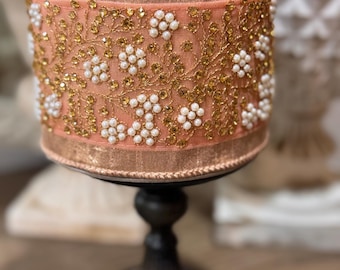 May include: A peach-colored ribbon with gold and white embellishments is wrapped around a black pedestal. The ribbon has a floral design with small white pearls and gold glitter.