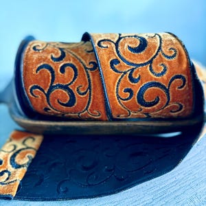 May include: A roll of wired ribbon featuring a plush, golden-brown velvet texture with a dark navy blue embroidered scrollwork pattern. The ribbon has a solid dark navy blue underside with a subtle, embossed matching scrollwork design throughout.