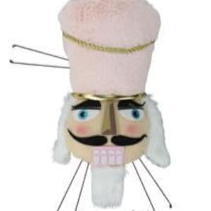 May include: A whimsical, plush nutcracker head with a pink, fuzzy hat and a gold-colored trim. The nutcracker has a tan face, blue eyes, a black mustache, and a white beard. It features wire attachments for hanging, perfect for holiday decor.