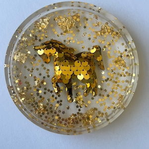 May include: A clear round coaster with a gold glitter background and a gold sequin unicorn in the center.