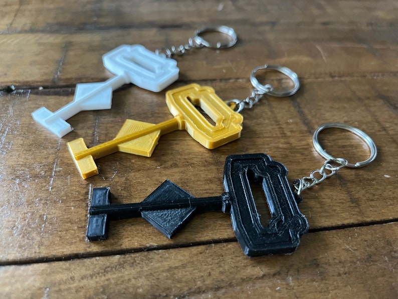May include: Three key-shaped keychains in black, gold, and white. Each keychain features a unique design with a keyhole and a diamond-shaped handle.