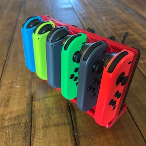 Puede incluir: Un conjunto de cinco mandos Nintendo Switch Joy-Con de colores en un soporte. Los mandos son azul, verde, amarillo, negro y rojo.
