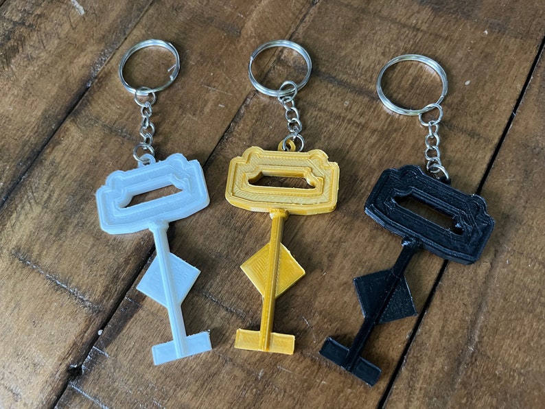 May include: Three key-shaped keychains in white, gold, and black. Each keychain has a unique design and is attached to a silver metal ring.