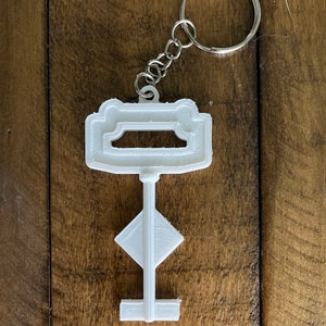 May include: White plastic key-shaped keychain with a silver metal ring. The key has a rectangular top with a cutout and a diamond-shaped middle section.