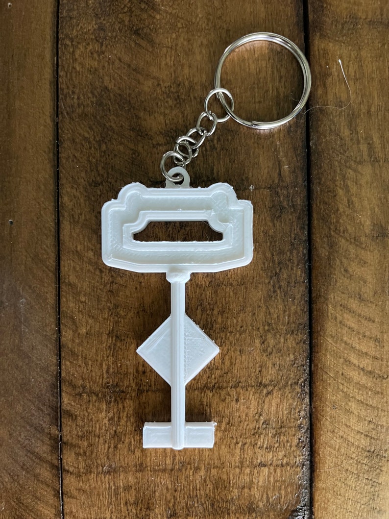 May include: White plastic key-shaped keychain with a silver metal ring. The key has a rectangular top with a cutout and a diamond-shaped middle section.