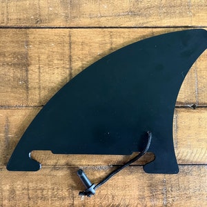 May include: Black surfboard fin with a black cord and a black plastic clip. The fin is on a wooden surface.
