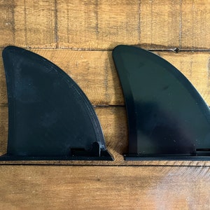 May include: Two black surfboard fins, also known as surf fins, are shown on a wooden surface. The fins are made of a hard plastic material and have a curved shape.