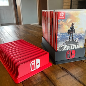 Puede incluir: Un soporte de tarjetas de juego de Nintendo Switch de plástico rojo con el logotipo de Nintendo Switch. El soporte tiene 12 ranuras para tarjetas de juego. Un soporte de tarjetas de juego de plástico negro con un logotipo de Nintendo Switch rojo sostiene una pila de estuches de juegos de Nintendo Switch. Los estuches de juegos presentan el juego "The Legend of Zelda: Breath of the Wild".