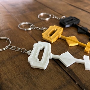 May include: Three keychains with different colored 3D printed designs. The keychain on the left is white, the middle keychain is gold, and the keychain on the right is black.
