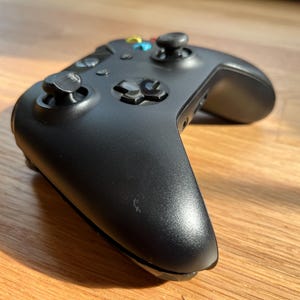 以下が含まれることがあります： 複数のボタンと2つのジョイスティックを備えた黒いゲームコントローラー。 コントローラーはマット仕上げで、木製の表面に置かれています。 ボタンの色は青、黄、赤です。