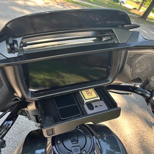 May include: Black motorcycle dashboard with a large screen, speakers, and a storage compartment. The compartment contains a phone, a lighter, and other small items. The motorcycle is parked on a street.