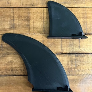 Puede incluir: Dos aletas de surf negras con una superficie texturizada. Las aletas están diseñadas para ser fijadas a una tabla de surf.