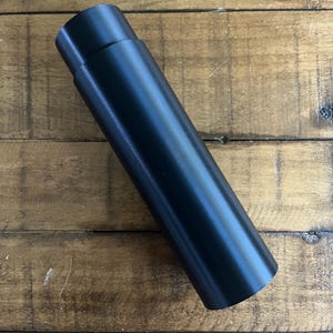 以下が含まれることがあります： 滑らかでマットな仕上がりの、円筒形の黒い金属製の物体。物体は段付きのデザインで、一端にわずかに狭いセクションがあります。アイテムは木製の表面に対して斜めに配置されています。