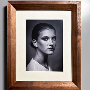Pode incluir: Retrato a preto e branco emoldurado em madeira. O retrato apresenta uma pessoa com cabelo escuro e uma blusa branca, com um passe-partout creme.