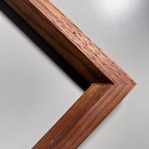 Op de afbeelding: Close-up van een houten fotolijsthoek. De lijst is een rijk, donkerbruin met zichtbare houtnerf. De hoek is precies verbonden, wat het vakmanschap laat zien. De achtergrond is zachtgrijs en contrasteert met de lijst.