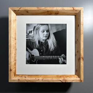 May include: A framed black and white photograph of a person playing an acoustic guitar. The photograph is set within a white mat and a light-colored wooden frame. The frame has a natural wood grain pattern.