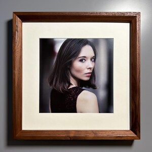 May include: A framed portrait of a woman with dark hair and a serious expression. The photograph is set within a cream-colored mat and a dark brown wooden frame. The woman is wearing a dark dress with a lace pattern.