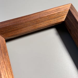 Peut inclure: Gros plan sur un coin de cadre en bois. Le cadre est en bois brun foncé avec un grain visible. Le cadre est incliné, montrant le joint d'angle et le bord intérieur. Le fond est gris clair.