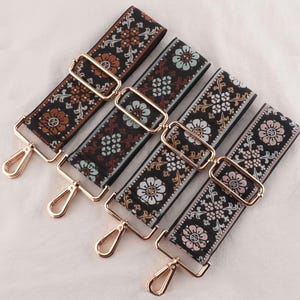 May include: Four embroidered bag straps with floral patterns in brown, blue, and pink on a black base. Each strap features gold-coloured metal hardware, including adjustable buckles and swivel clasps for attachment.