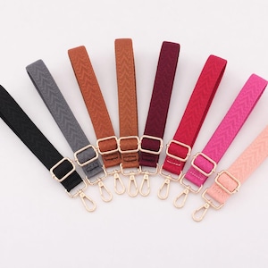 Pode incluir: Um conjunto de oito alças de bolsa ajustáveis dispostas em leque. Cada alça apresenta um padrão de espinha de peixe tecido e ferragens de metal dourado, incluindo uma fivela deslizante e um mosquetão. As cores incluem preto, cinza, castanho claro, castanho, bordô, vermelho, magenta e rosa claro.