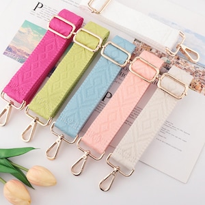 May include: A collection of colorful, patterned bag straps in shades of pink, green, blue, peach, and white. Each strap features gold-colored metal hardware, including adjustable buckles and swivel clasps. The straps are arranged on a printed paper background.