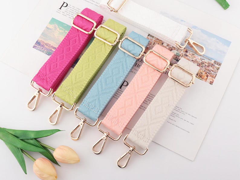 May include: A collection of colorful, patterned bag straps in shades of pink, green, blue, peach, and white. Each strap features gold-colored metal hardware, including adjustable buckles and swivel clasps. The straps are arranged on a printed paper background.