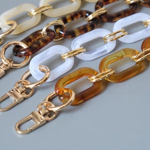 May include: Four bag straps featuring interlocking rectangular acrylic links in cream, tortoiseshell, white, and translucent amber. Each strap is finished with a polished gold-tone metal lobster clasp at the end for secure attachment to a handbag.