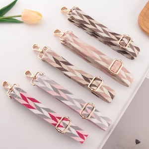 May include: Six patterned bag straps in various colours, including pink, grey, and brown, with gold-coloured metal hardware. The straps feature a geometric zigzag design. A tulip and a magazine are in the background.