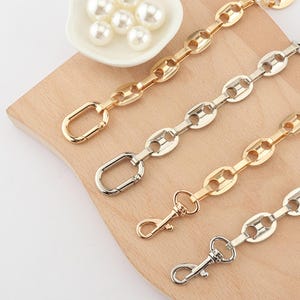 May include: Close-up of several metal chain straps in gold and silver tones, with oval links and lobster claw clasps. The chains are arranged on a light wooden surface, with a small dish of white pearls in the background.