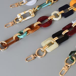 Può includere: Una collezione di cinturini a catena in acrilico colorato con chiusure in metallo dorato. Le catene presentano varie forme e colori, tra cui maglie rettangolari, ovali ed esagonali nelle tonalità del marrone, blu, rosso, nero e bianco.