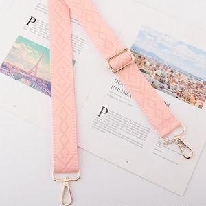 May include: A light pink bag strap with a geometric pattern and gold-colored metal hardware. The strap is lying on a magazine with images of a city and the Eiffel Tower.